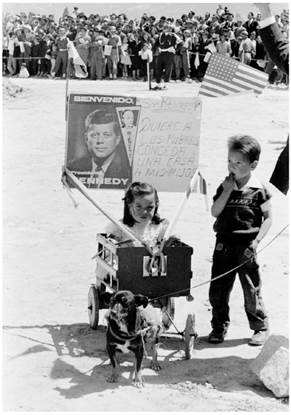 Una niña (sentada en una carreta) y un niño asisten a la ceremonia de colocación de la primera piedra en el Proyecto de Vivienda de Ciudad Techo, en Bogotá; delante de la carreta hay un perro pequeño y en la parte trasera de la carreta se ve un cartel escrito en español que dice “Sr. Kennedy. Quiere a los pobres conceda una casa a mis hijos”, 17 de diciembre de 1961